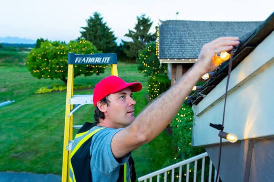 An installer hanging Christmas lights along a roofline from a ladder